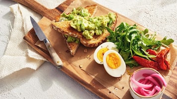 A wooden cutting board with toast, avocado, eggs, greens, and pickles.