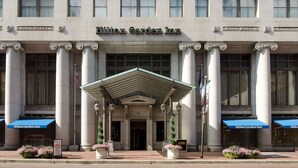 El exterior del Hilton Garden Inn, con un diseño arquitectónico clásico, presenta grandes columnas, una entrada destacada y un toldo azul.