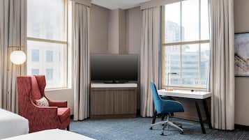 A hotel room with a bed, a desk with a TV, a red armchair, a blue office chair, and large windows with curtains.