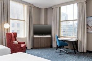A hotel room with a bed, a desk with a TV, a red armchair, a blue office chair, and large windows with curtains. - Hilton Garden Inn Indianapolis Downtown (Indianapolis)