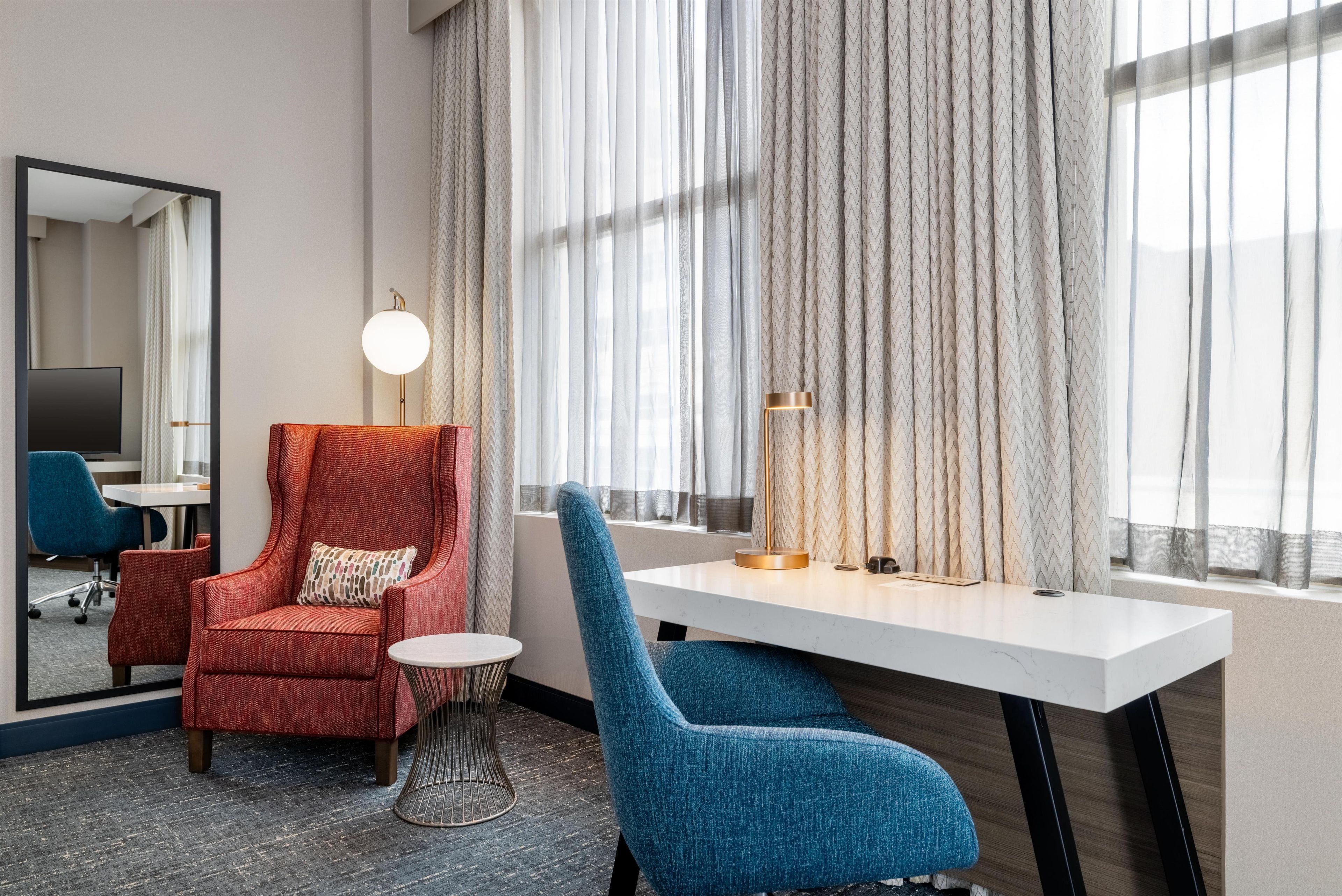 Una habitación de hotel con un sillón rojo, un sillón azul, una mesita redonda pequeña, un escritorio, una lámpara, un espejo y cortinas de encaje.