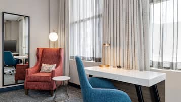 Una habitación de hotel con un sillón rojo, un sillón azul, una mesita redonda pequeña, un escritorio, una lámpara, un espejo y cortinas de encaje.