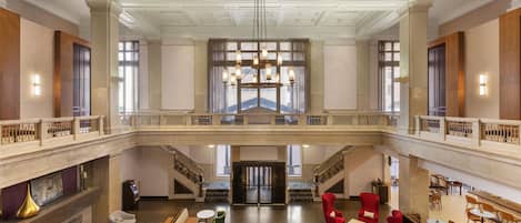 A spacious lobby with a high ceiling, a grand staircase, and a seating area with red chairs and round tables.