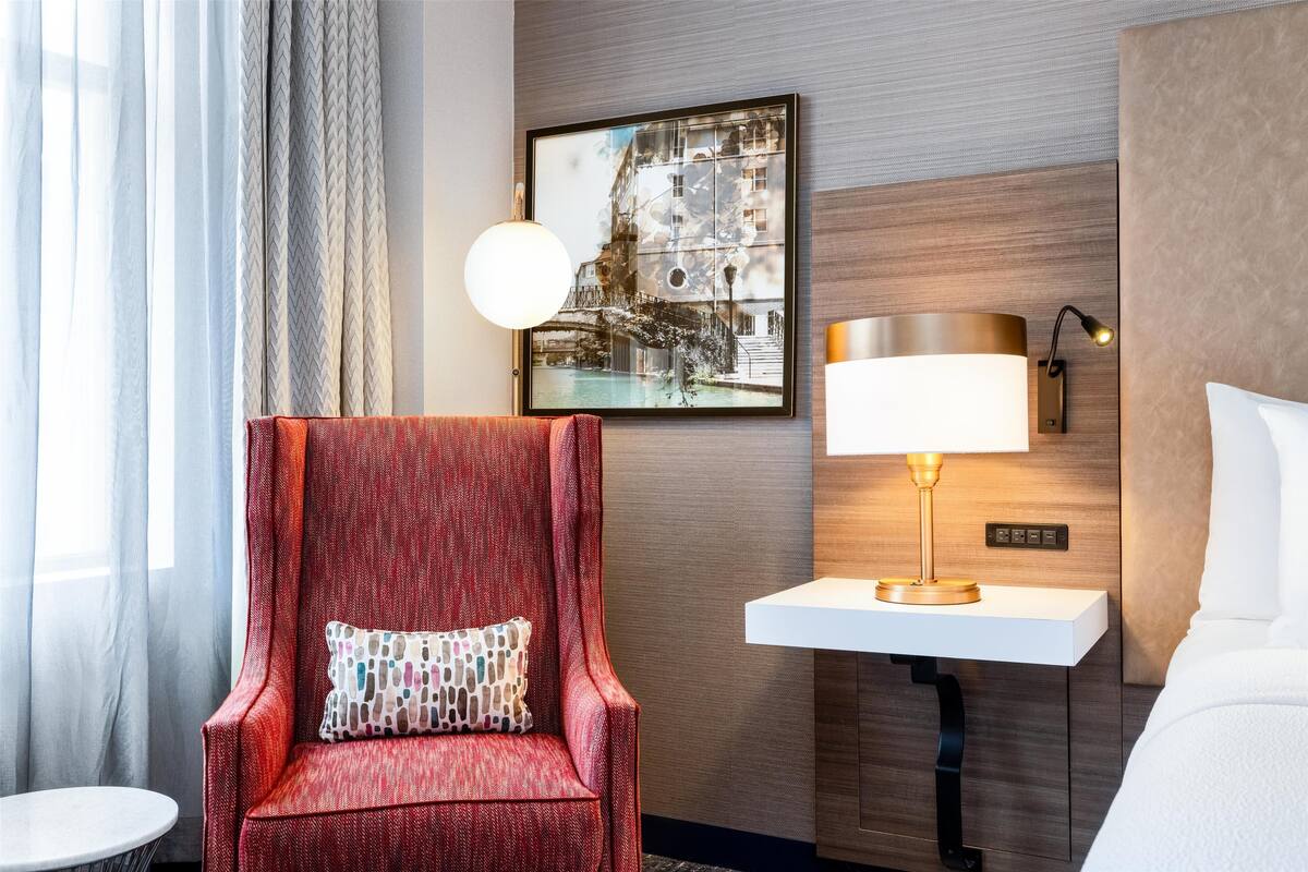 Ein Hotelzimmer mit einem roten Sessel, einem weißen Nachttisch mit Lampe, einem Stadtbild im Bilderrahmen und einem Bett mit weißer Bettwäsche.