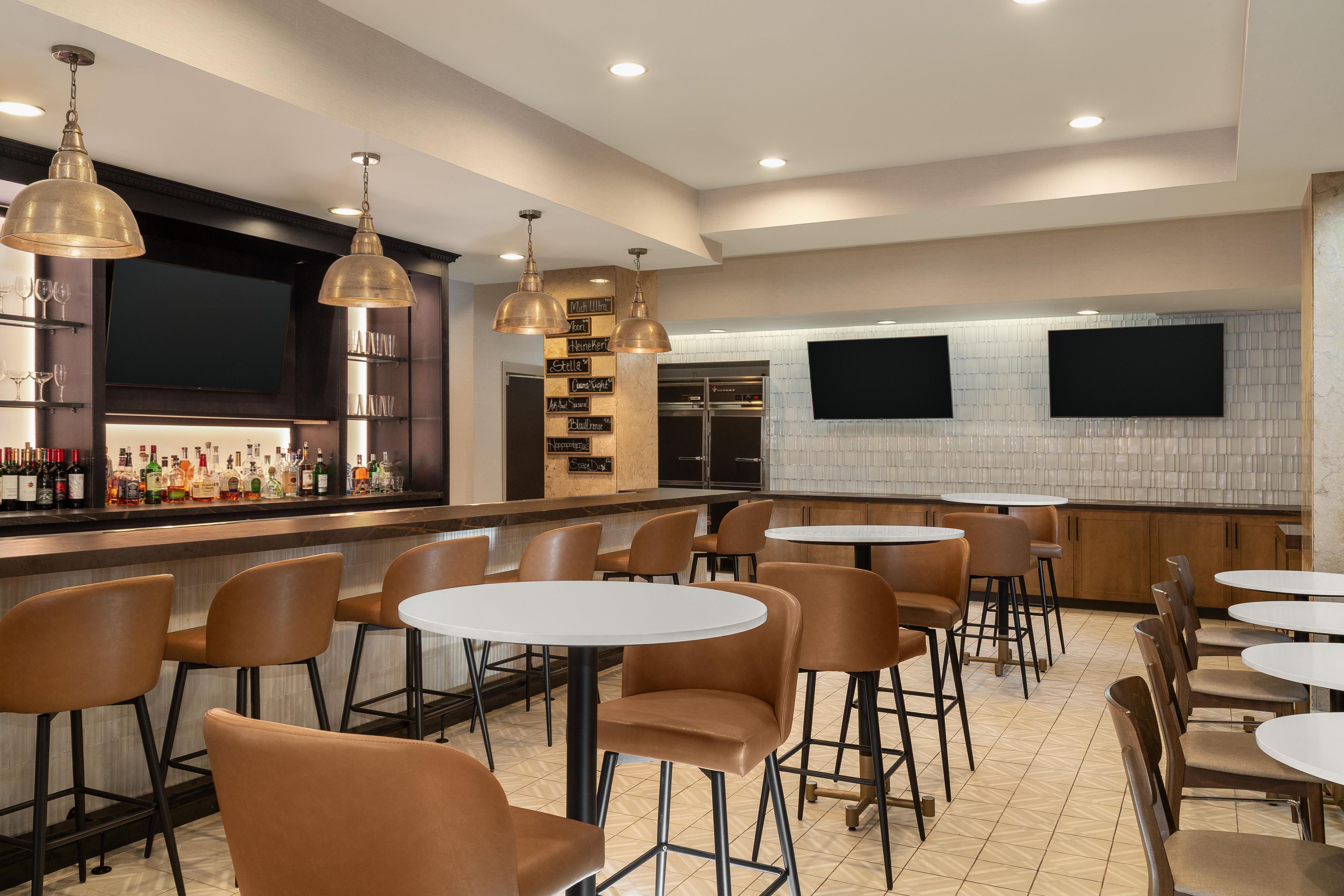 A bar area with multiple seating options, including round tables and chairs, and a selection of bottles on the shelves.