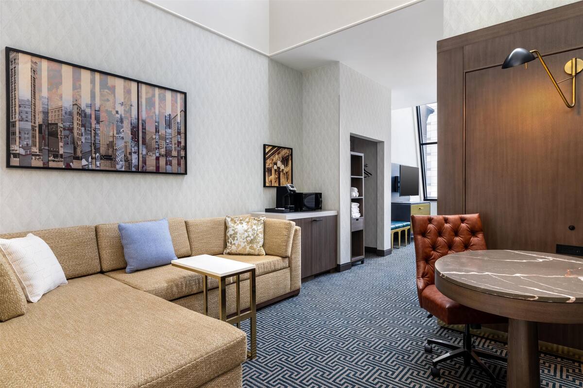 Ein Hotelzimmer mit einer Eckcouch, einem runden Tisch, einem Essbereich und einem Stadtbild an der Wand.