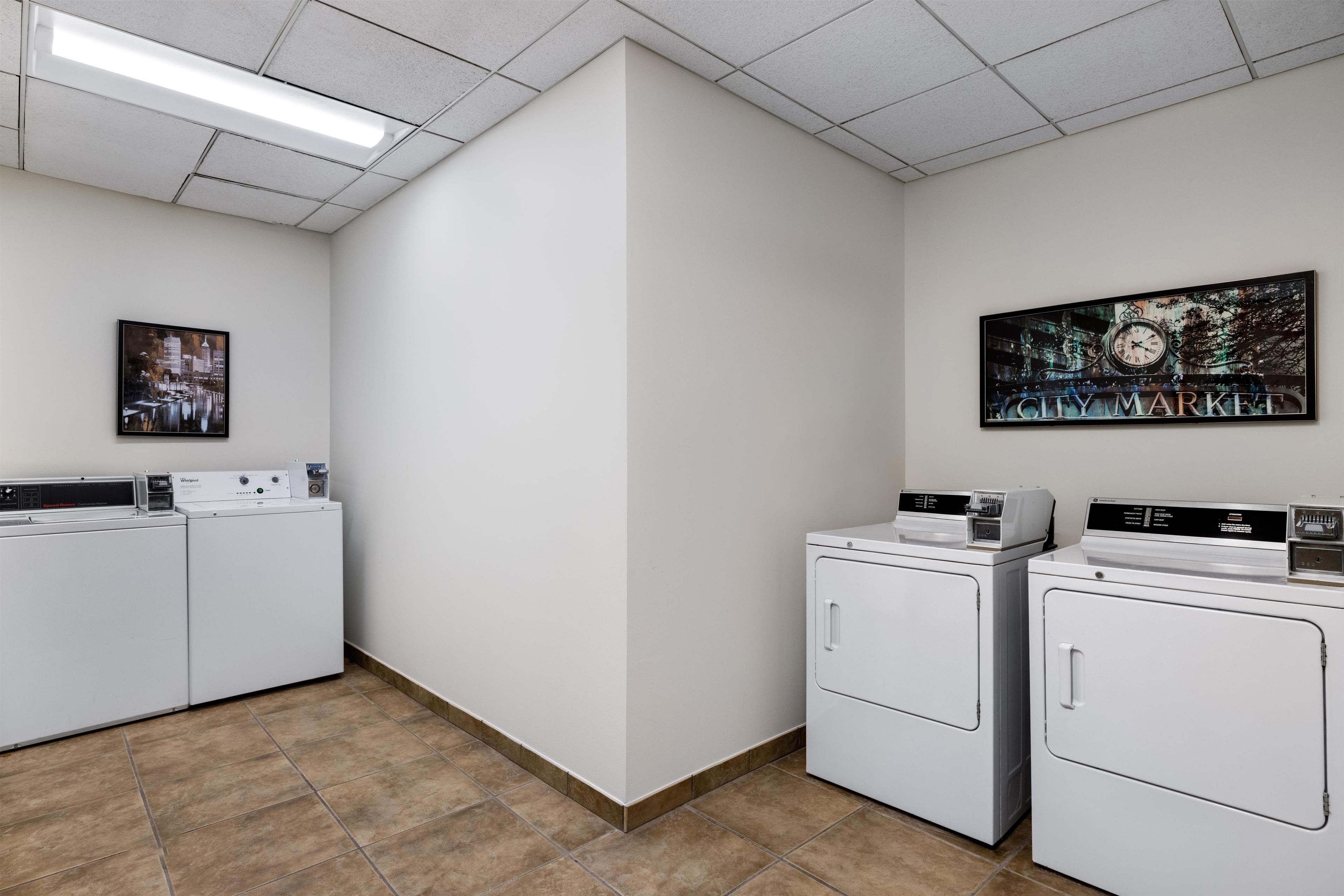 Een wasserette met vier witgekleurde voorladers, wasmachines en drogers, een groot stadsbeeld in een lijst aan de muur en een fluorescentiearmatuur aan het plafond.