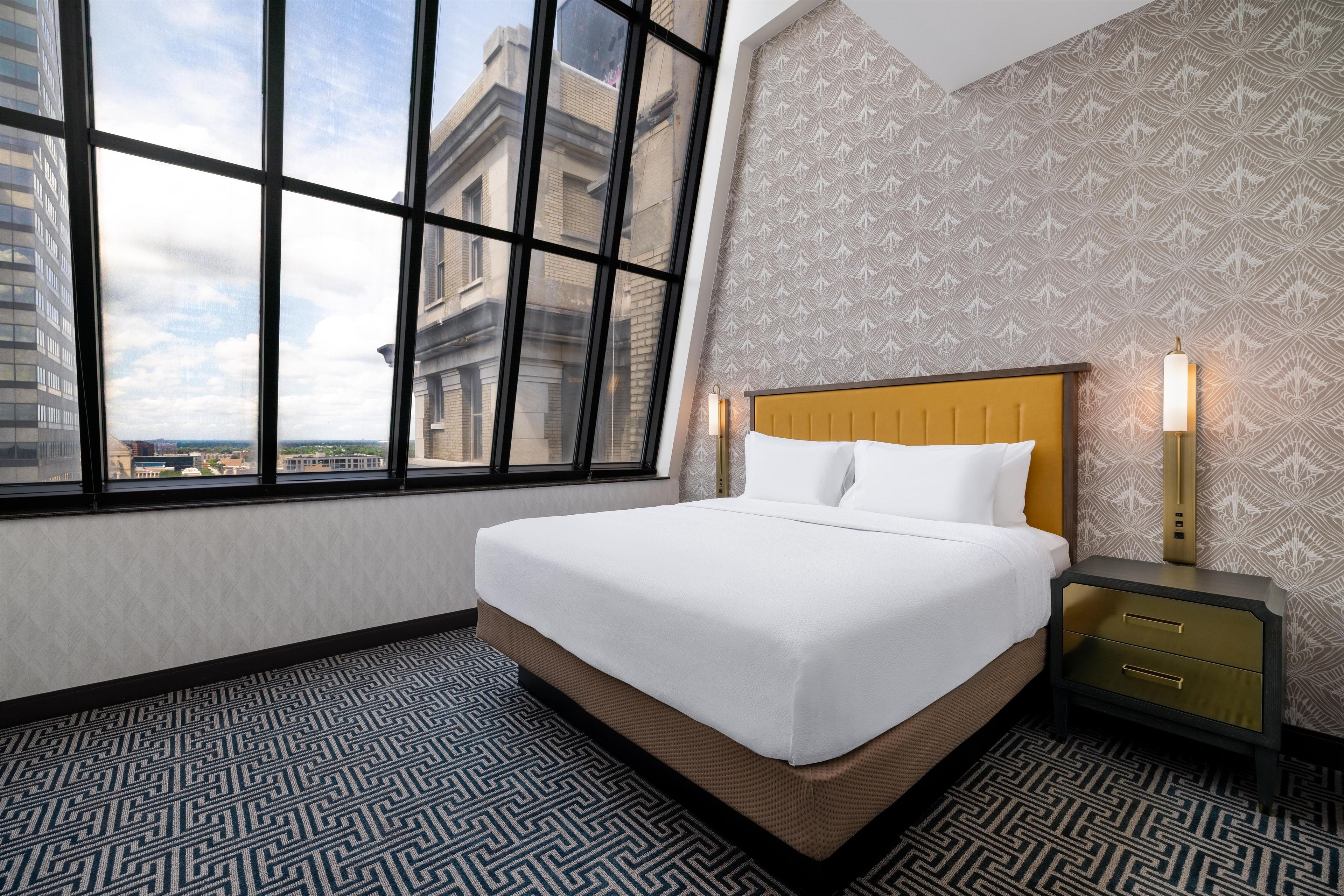 A hotel room with a large window, a bed with white linens, a nightstand with a lamp, and patterned wallpaper.