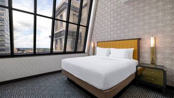 A hotel room with a large window, a bed with white linens, a nightstand with a lamp, and patterned wallpaper.