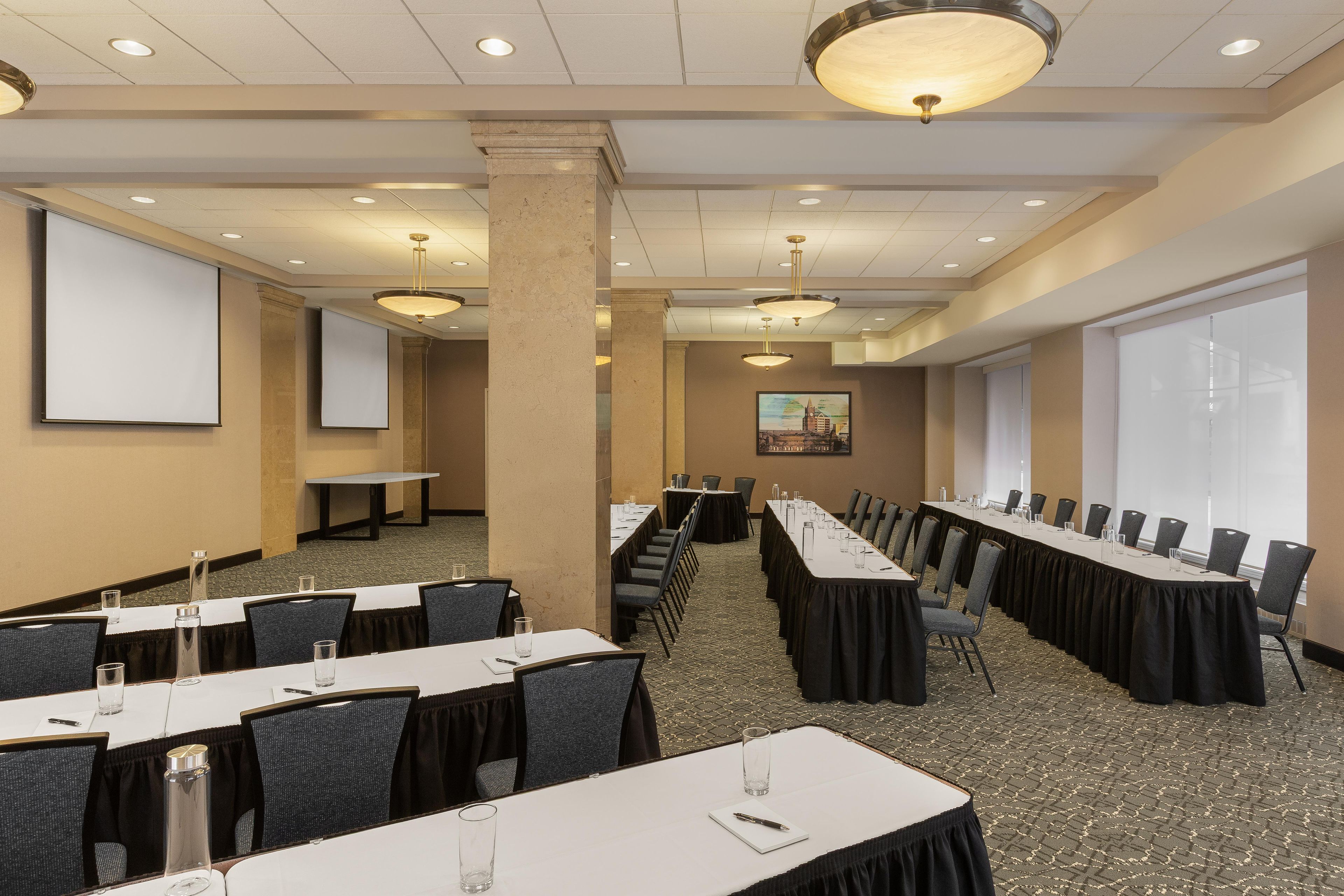 A conference room with long tables, chairs, a projector screen, and a large window.