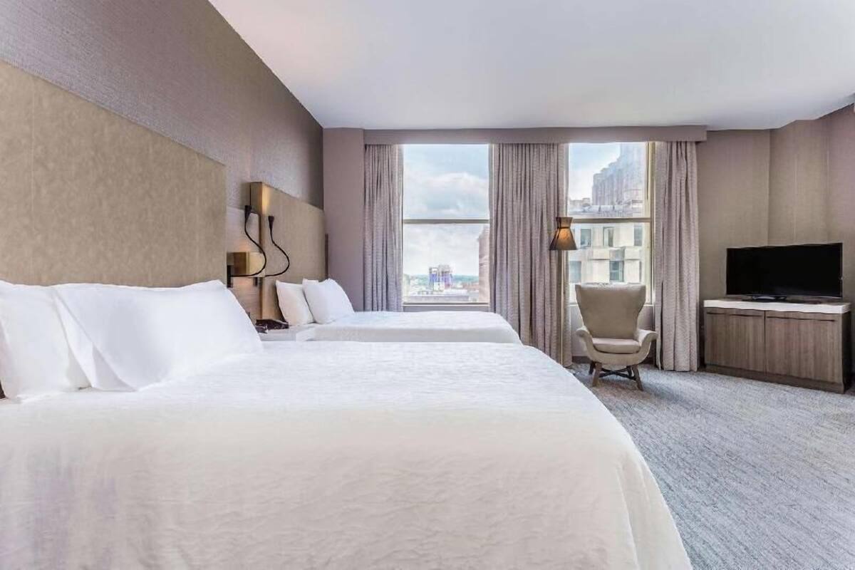 Ein Hotelzimmer mit zwei Betten, einem Fernseher, einem Sessel und Blick auf eine Stadtlandschaft.