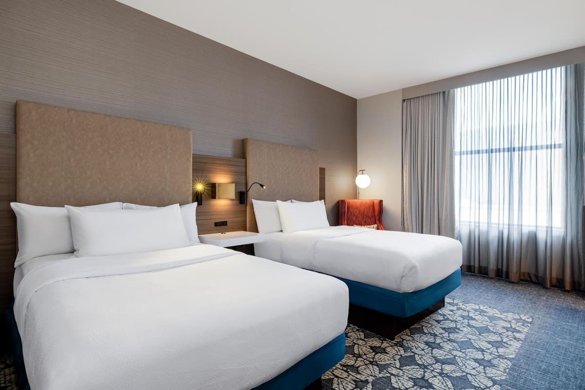 Ein Hotelzimmer mit zwei Betten, einem großen Fenster mit Vorhängen, einem gemusterten Teppich und einer Wandleuchte.