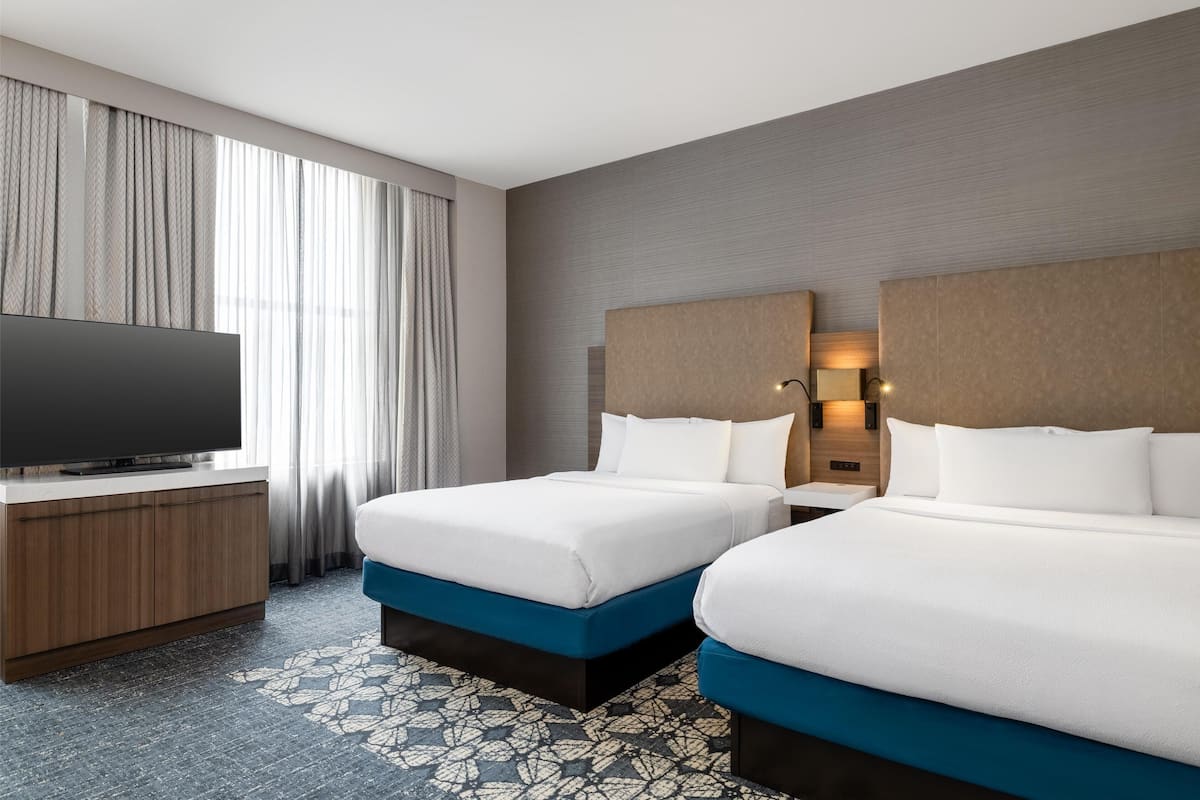 Ein Hotelzimmer mit zwei Betten, einem Fernseher, einer Holzkommode und einem gemusterten Teppich.