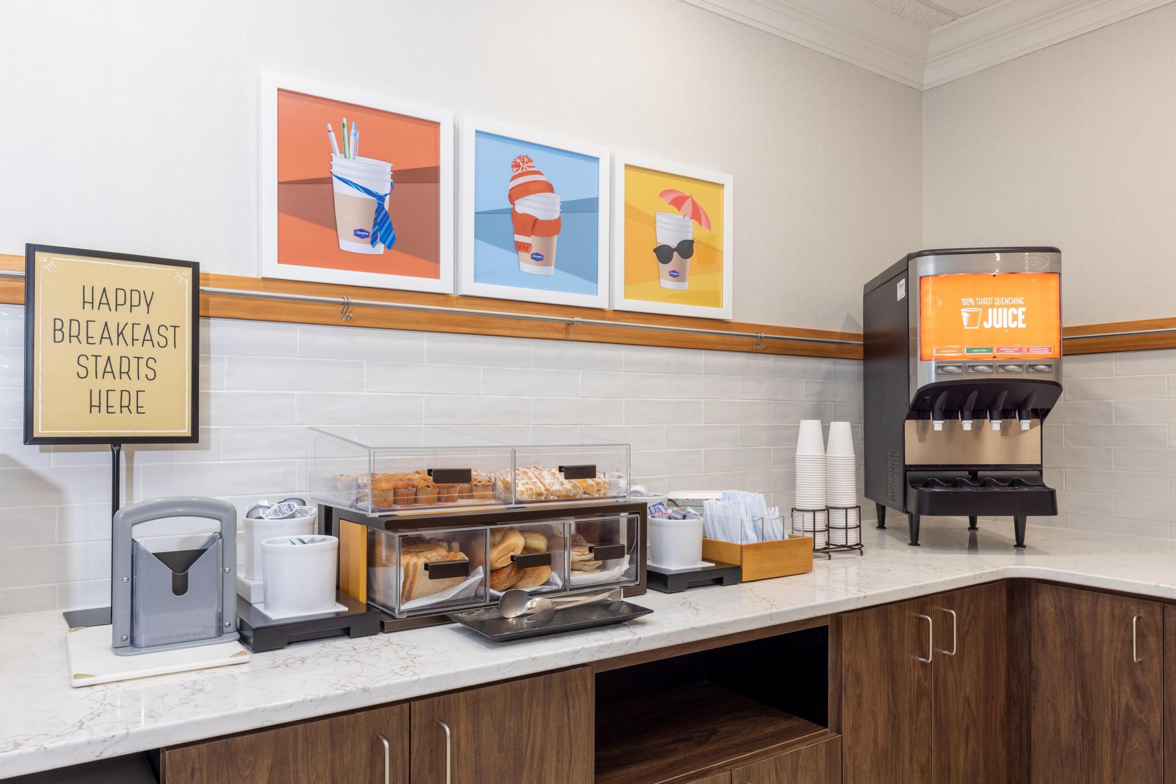 A breakfast station with a juice dispenser, a sign that says 'HAPPY BREAKFAST STARTS HERE,' and a display of pastries.