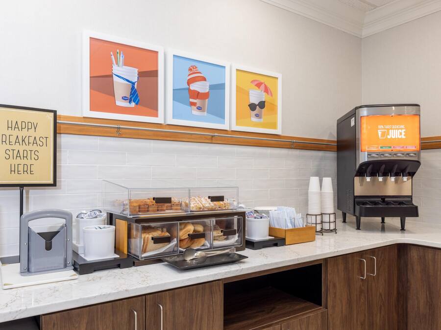 A breakfast station with a juice dispenser, a sign that says 'HAPPY BREAKFAST STARTS HERE,' and a display of pastries.