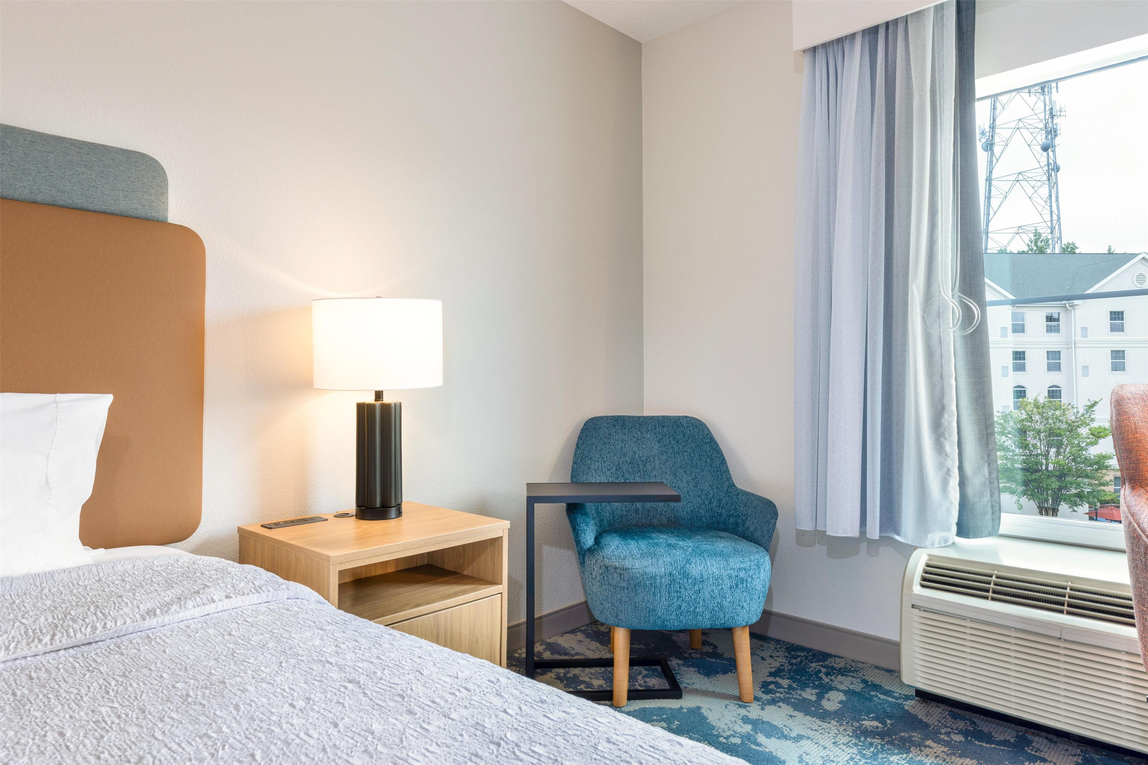 A hotel room with a bed, a desk with a lamp, a blue armchair, a window with curtains, and an air conditioner.