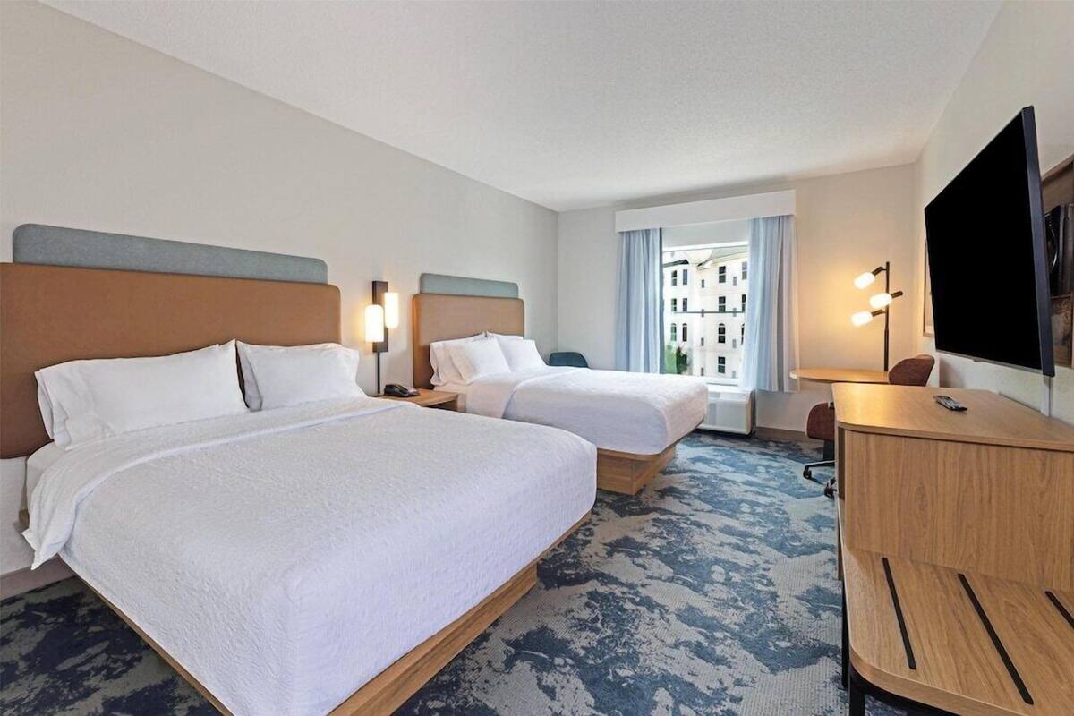 A hotel room with two beds, a desk, a TV, and a window with a view of buildings.