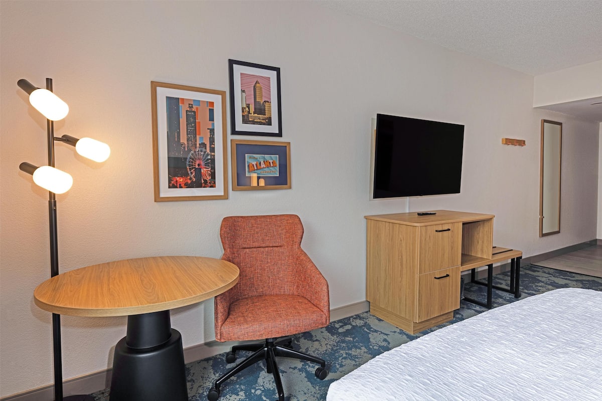 Una habitación de hotel con una mesa redonda de madera, una silla naranja, un televisor, un armario de madera y una cama.