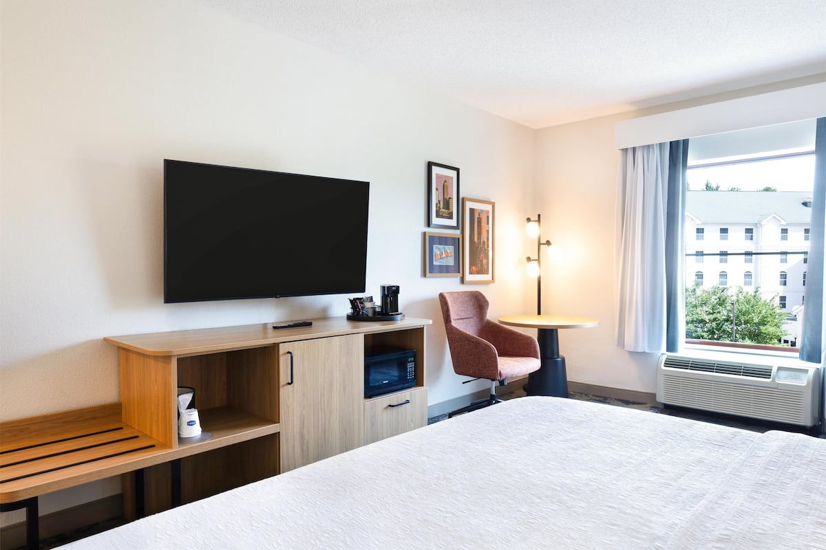 Habitación de hotel con cama, televisión, escritorio con cafetera, una silla y ventana con vista a edificios.