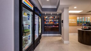 Un vestíbulo de hotel con un expositor refrigerado de bebidas, estantes con refrigerios, un mostrador de recepción y un cartel que dice 'Welcome to Hampton'.