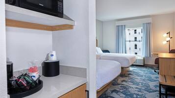 Habitación de hotel con microondas, cafetera y una pequeña mesa con una taza y pañuelos. Hay dos camas, una alfombra estampada azul y una ventana con cortinas.