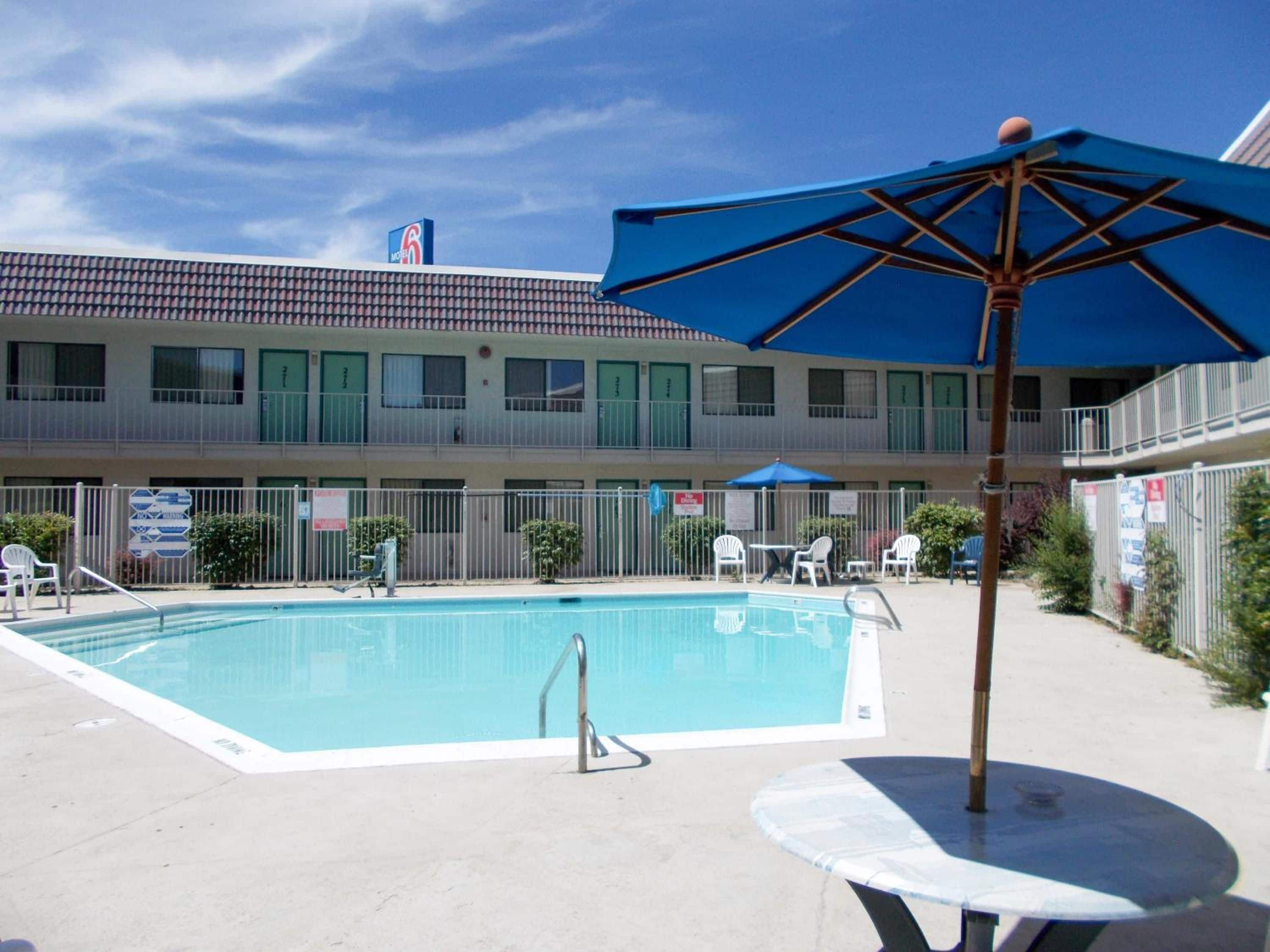 A swimming pool with a blue umbrella, surrounded by a fenced area with lounge chairs and a motel building in the background.