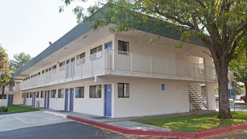 Un motel de dos pisos con un balcón en el nivel superior, un estacionamiento y un árbol en primer plano.