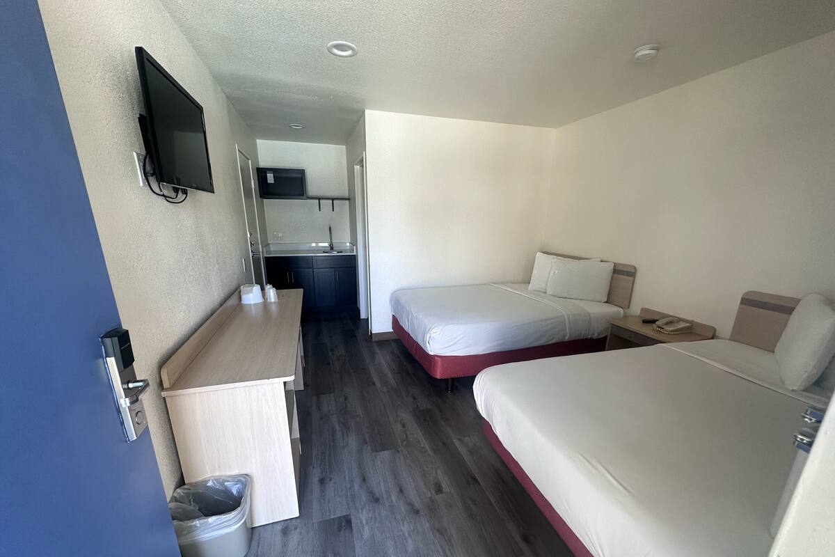 A hotel room with two beds, a small kitchenette, a TV mounted on the wall, and a desk with a trash bin.