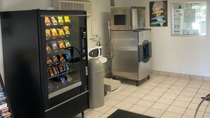Ein Getränkeautomat mit Snacks, eine Mikrowelle und ein Mülleimer in einem Raum mit gefliestem Boden und einer schwarzen Matte.