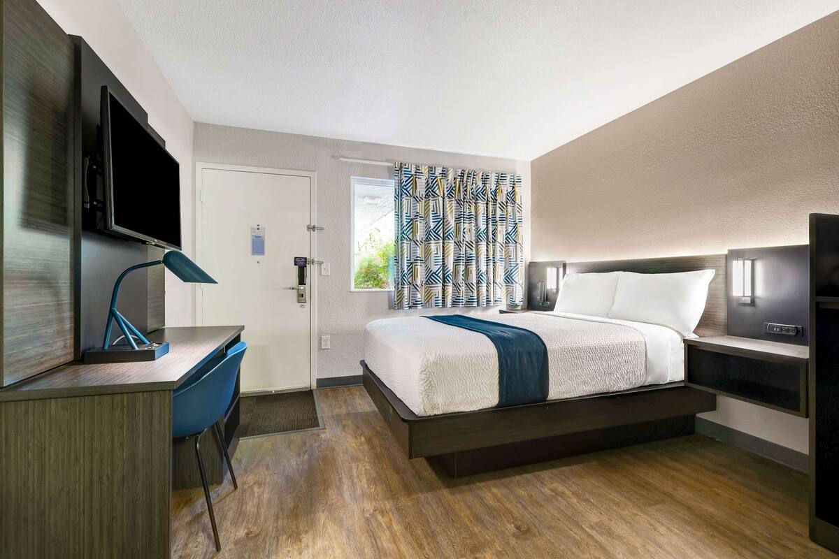 A modern hotel room with a bed, desk, chair, TV, and a window with patterned curtains.