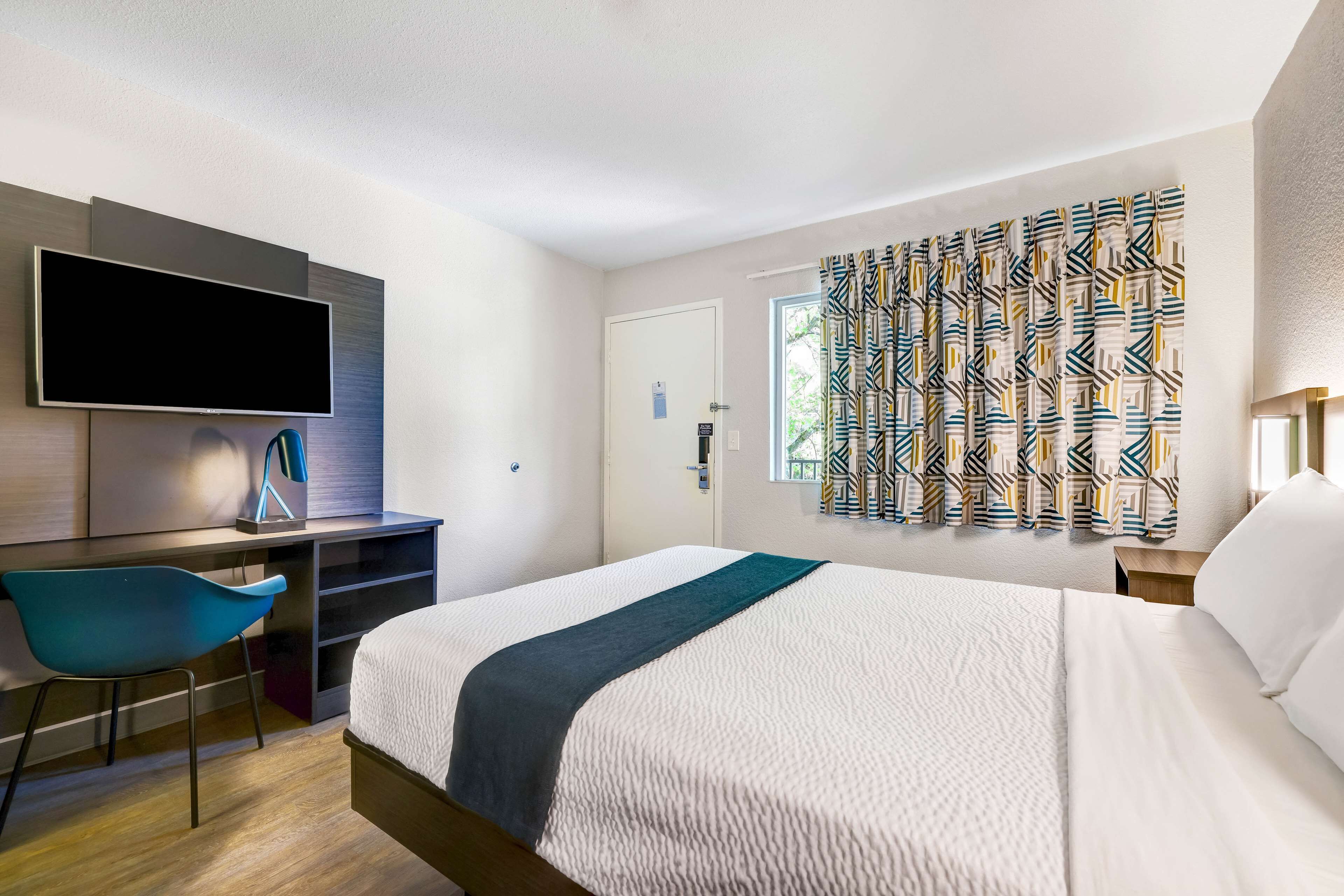 A hotel room with a bed, a desk with a TV, a chair, and a window with patterned curtains.