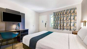 A hotel room with a bed, a desk with a TV, a chair, and a window with patterned curtains.