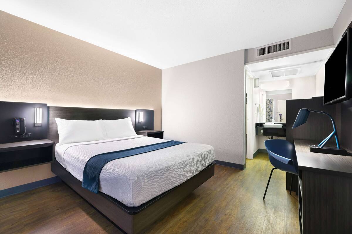 A modern hotel room with a bed, bedside table, and a desk with a chair.