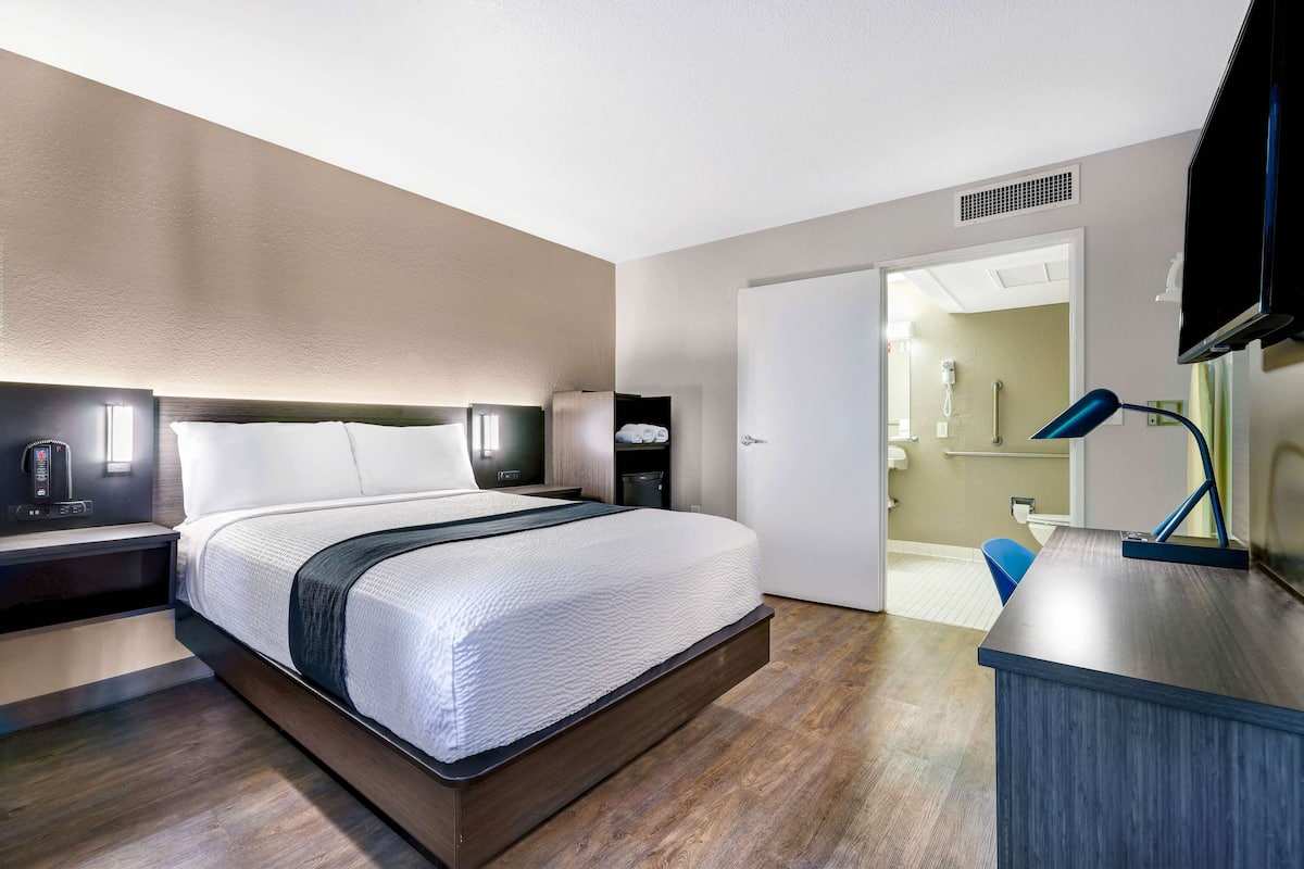 A modern hotel room with a large bed, bedside tables, a desk with a lamp, and a bathroom visible through an open door.