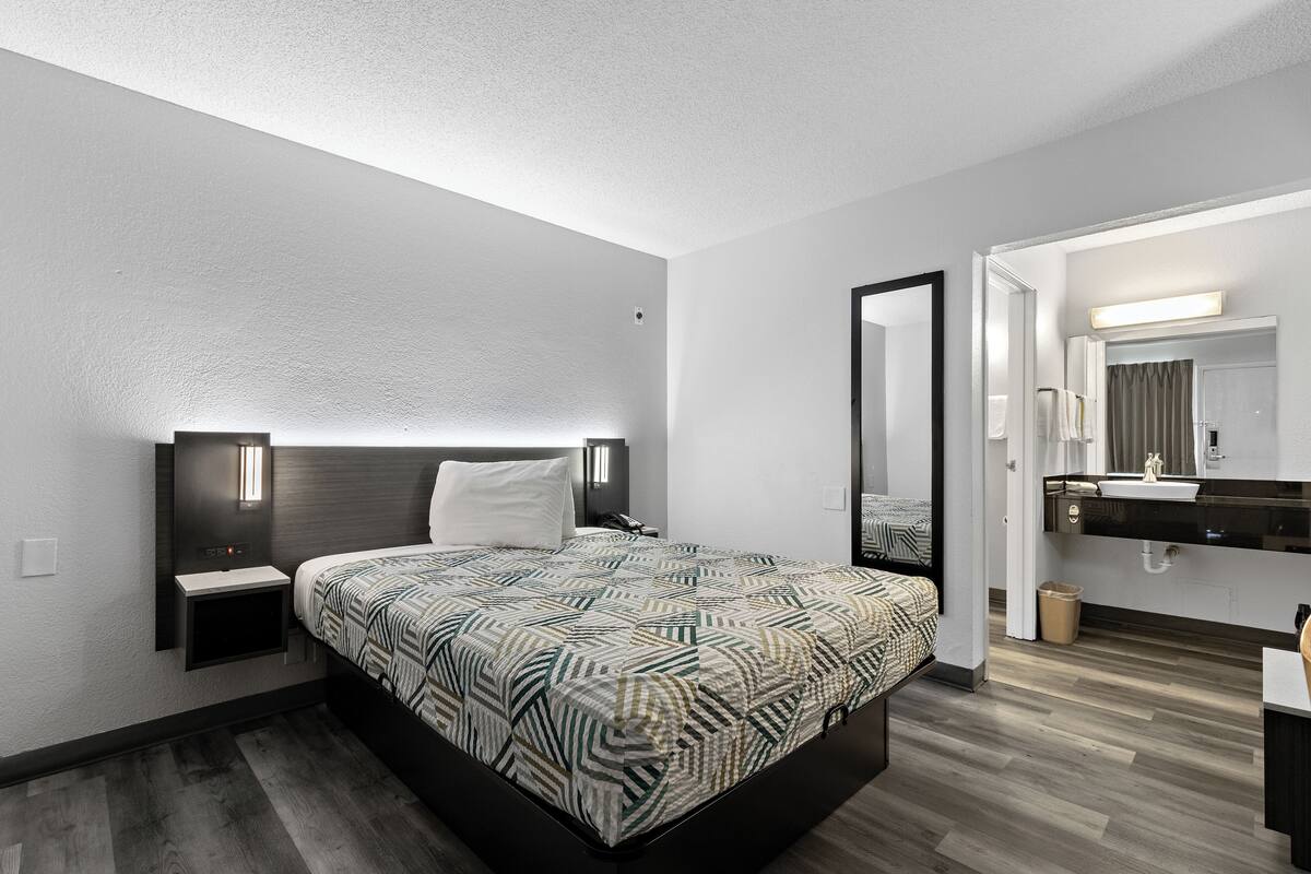 A modern hotel room with a large bed, bedside tables, a mirror, and a bathroom with a sink and mirror.