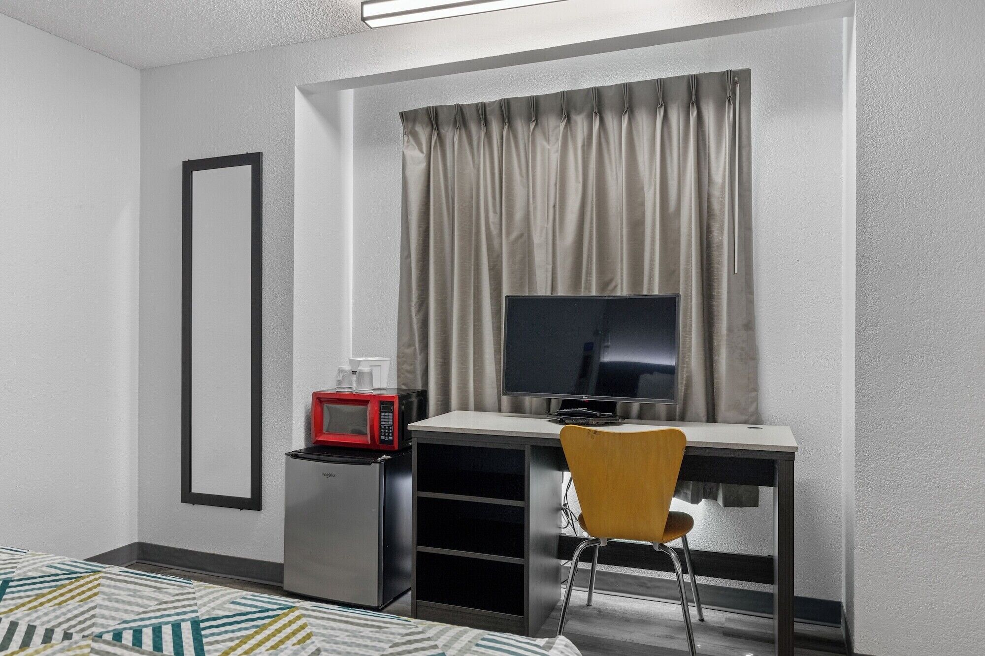 Ein kleines Hotelzimmer mit einem Bett, einem Schreibtisch mit Computer und Fernseher, einer kleinen Kühlbox und einem Sessel.