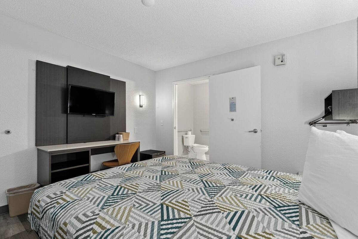 A hotel room with a bed, a desk with a flat-screen TV, a chair, a toilet, and a door.
