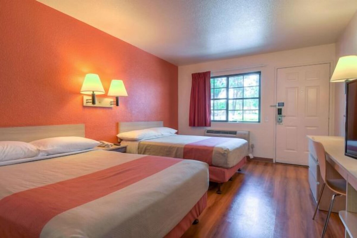 A hotel room with two beds, a desk, a chair, a window with curtains, and wall-mounted lamps.