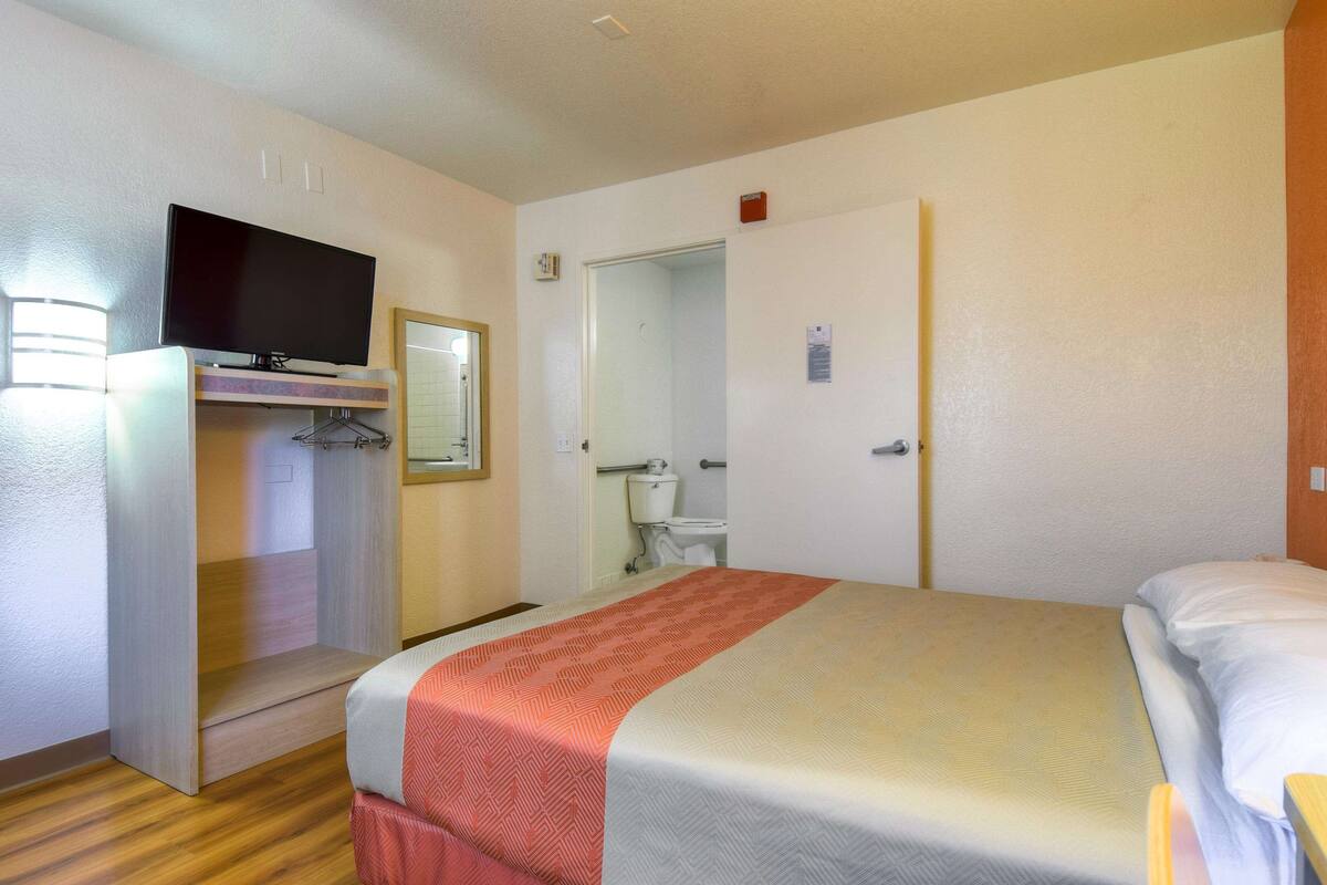 A hotel room with a bed, a television on a stand, a bathroom, and a door.
