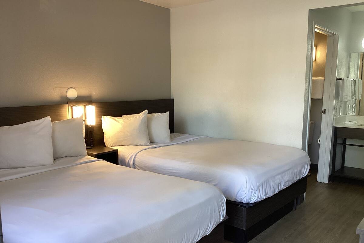 A hotel room with two beds, a nightstand with a lamp, a bathroom with a mirror, and a wooden floor.
