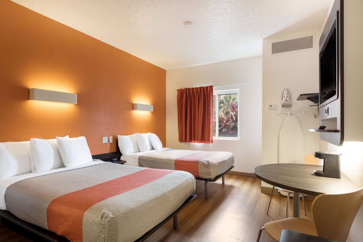 Ein Hotelzimmer mit zwei Betten, einem runden Esstisch, einem Fernseher und einem Fenster mit Vorhängen.