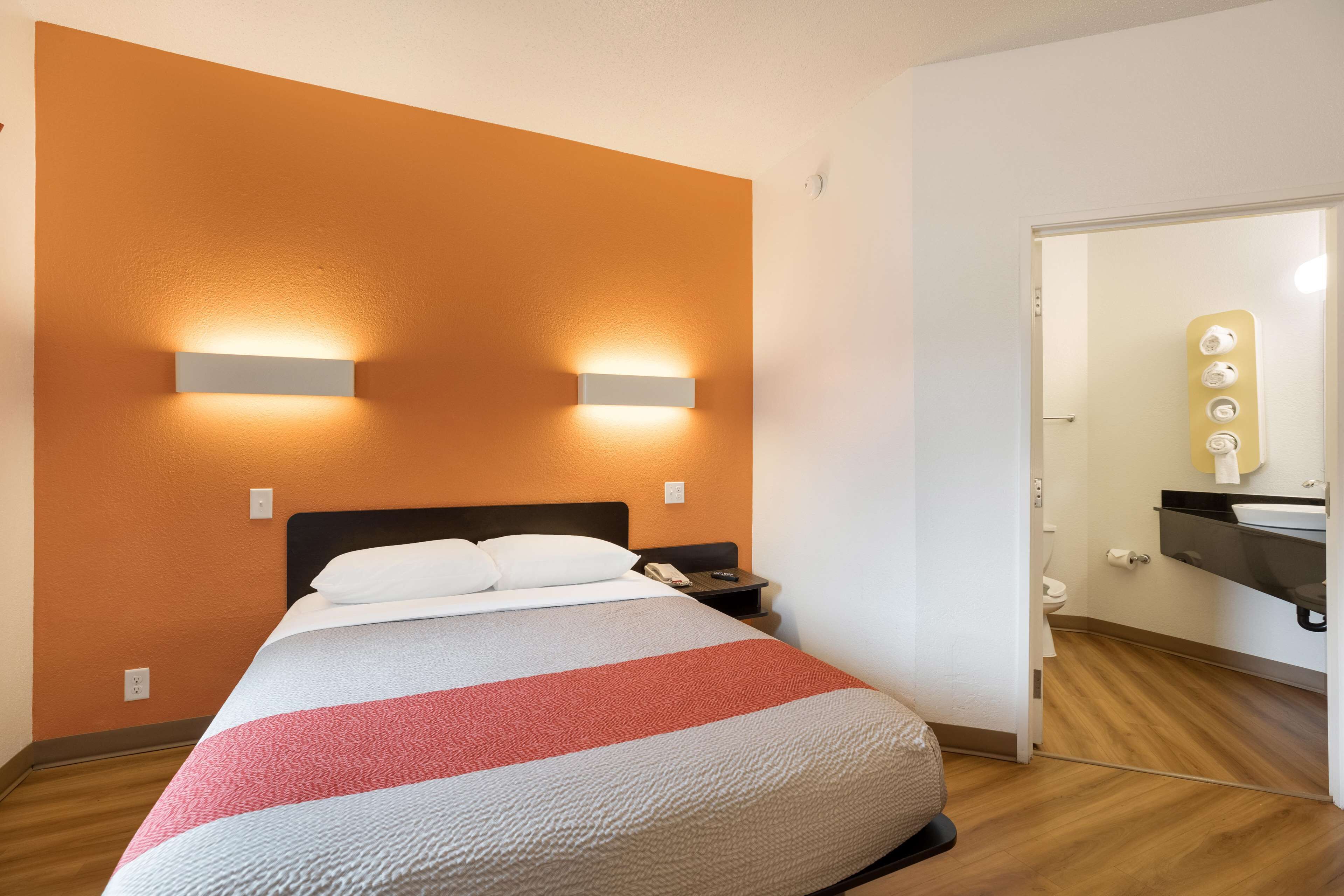 Habitación de hotel con cama, mesita de noche, lámpara en la pared y baño visible a través de una puerta abierta.