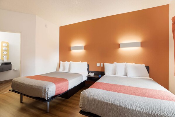 Una habitación de hotel con dos camas, piso de madera, lámpara de pared y mesita de noche con teléfono y lámpara.