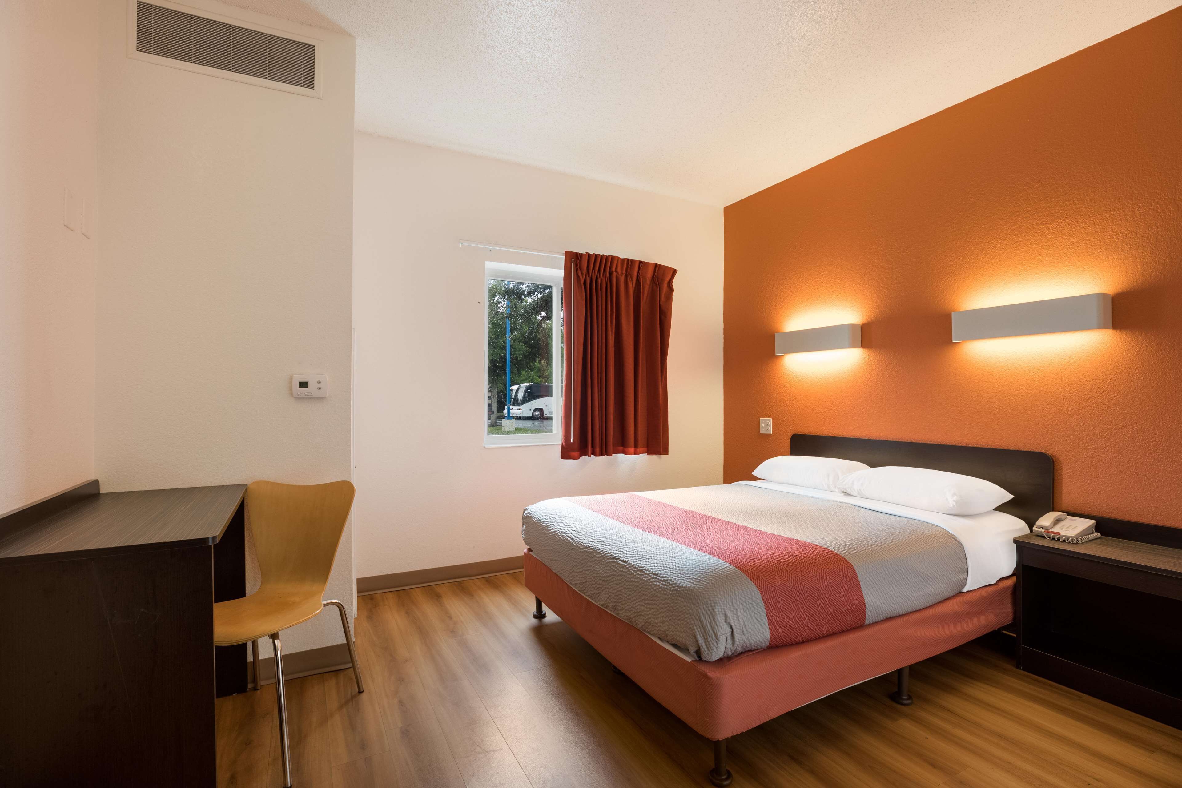 A hotel room with a bed, a desk, a chair, a window with curtains, and wall-mounted lights.