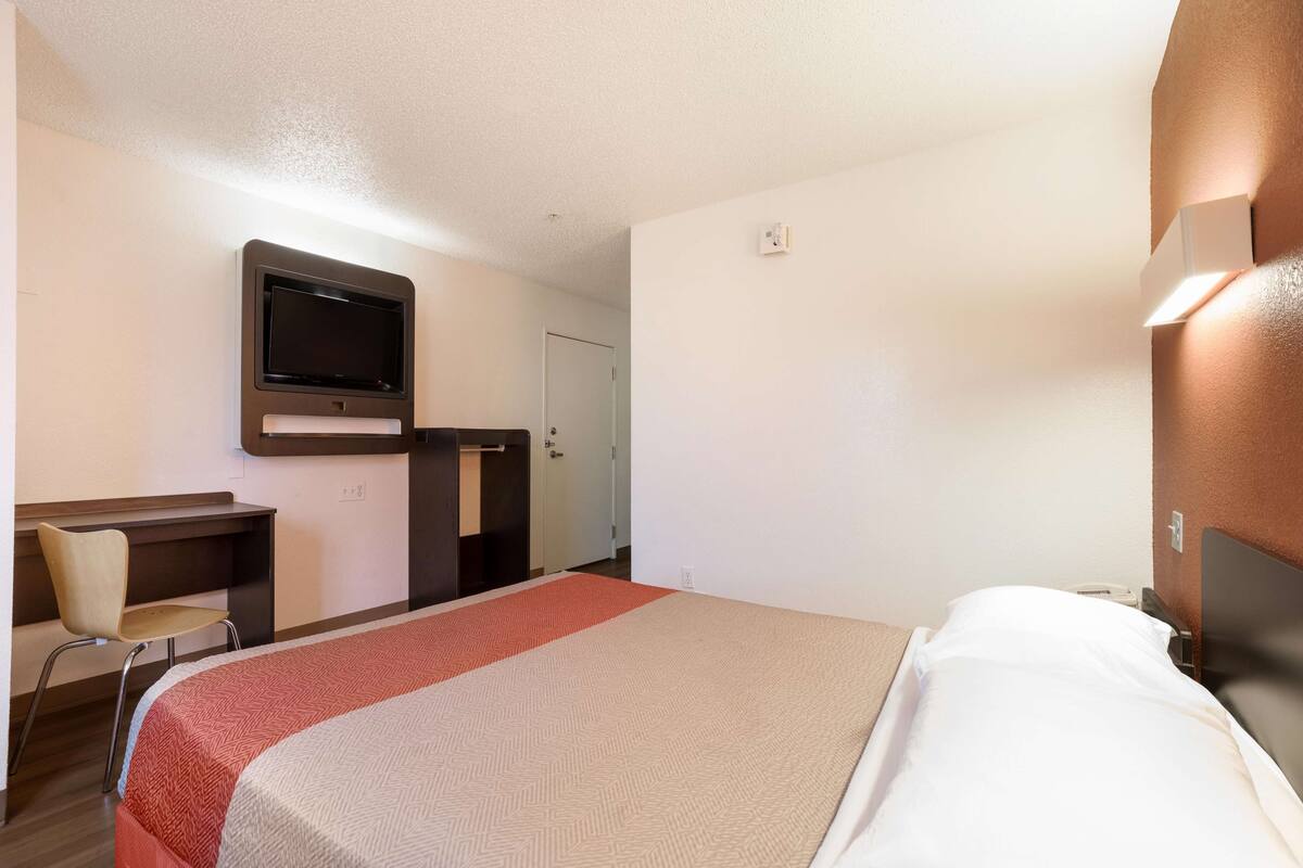 Ein Hotelzimmer mit Bett, Schreibtisch mit Stuhl, Wandfernseher und einer Tür zu einem anderen Raum.