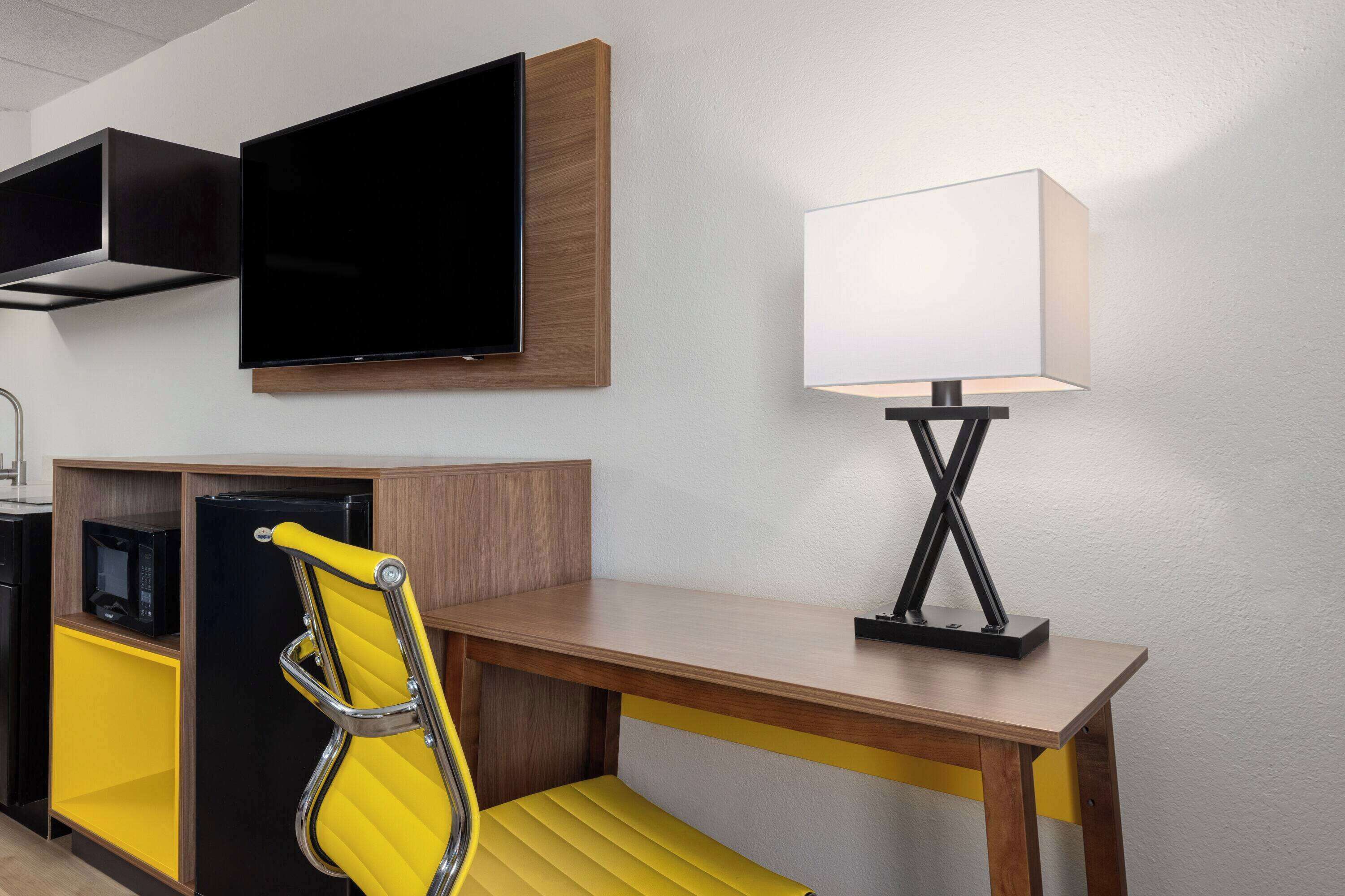 Une chambre d’hôtel moderne avec un bureau en bois, une chaise jaune, un téléviseur à écran plat, une lampe et un four à micro-ondes.