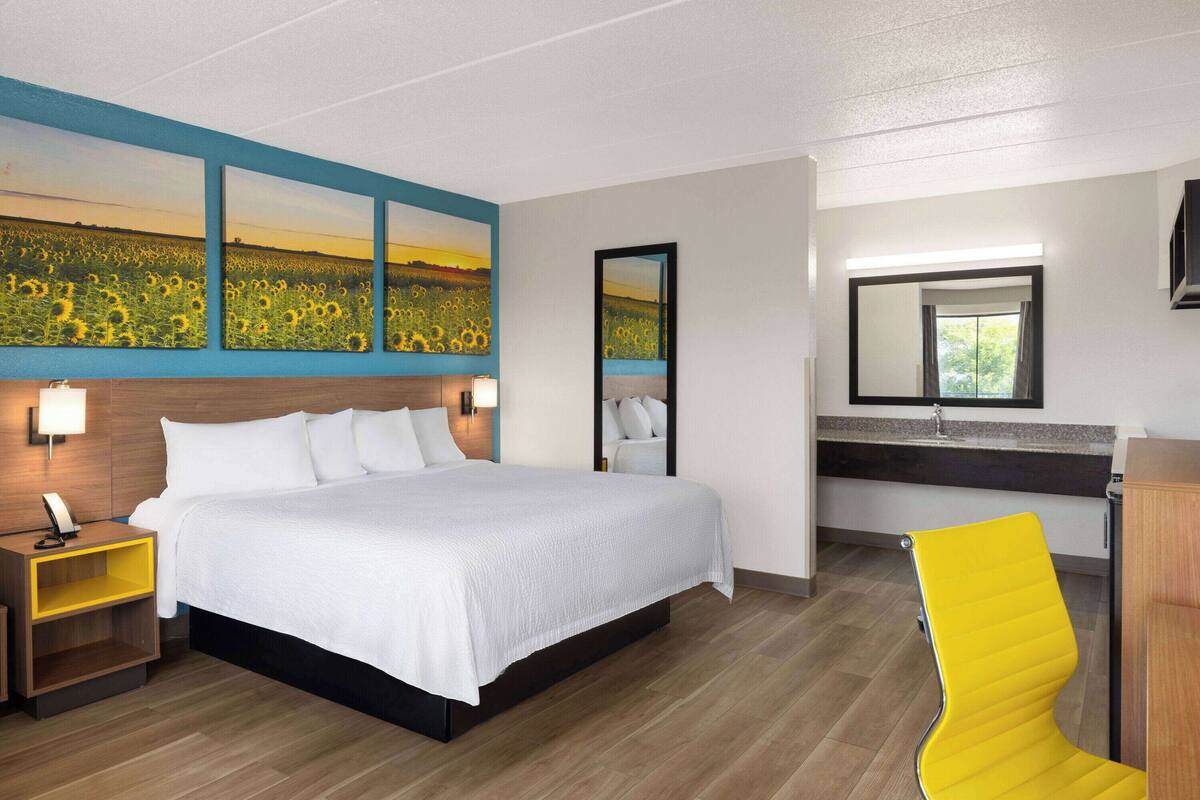 Una habitación de hotel con una cama grande, una mesita de noche con lámpara, un espejo, una silla amarilla y un cuadro de girasoles en la pared.