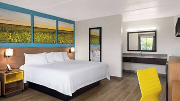 A hotel room with a large bed, a bedside table with a lamp, a mirror, a yellow chair, and a painting of sunflowers on the wall.