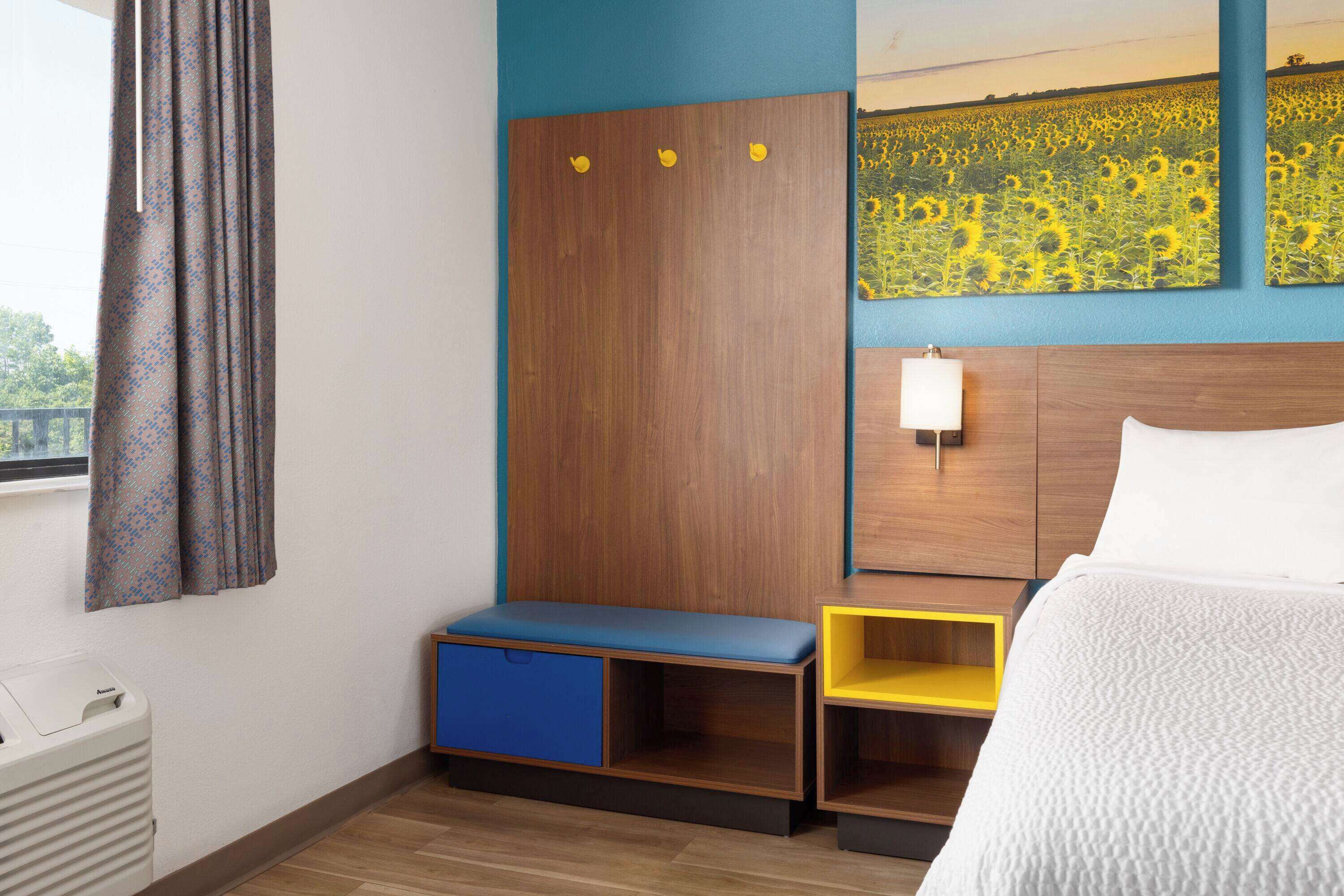 Ein Hotelzimmer mit einem Holz-Kopfteil, einer Sitzbank mit blauem und gelbem Stauraum, einem Nachttisch mit Lampe und einem großen Gemälde einer Sonnenblume an der Wand.