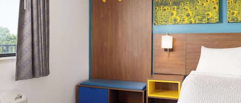 A hotel room with a wooden headboard, a bench with blue and yellow storage, a bedside table with a lamp, and a large sunflower painting on the wall.